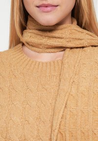 Soe villane kampsun sooja karamellitooniga, millel on palmikunähtus, kombineeritud sobiva tekstuuriga salliga, mis on ümber kaela mähitud.