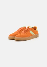 Oransje semsket sneakers med lys grønne detaljer, snøring, gummisåle og teksturert overflate. Har en stripe detalj på siden.