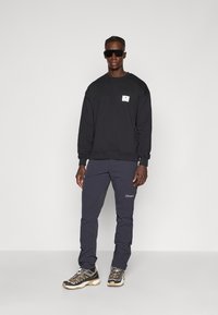 Felpa nera con patch logo, pantaloni navy con logo e scarpe da trekking multicolori con suole testurizzate. Gli occhiali da sole completano l'outfit.