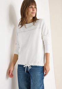 Witte longsleeve top met een ronde halslijn, een trekkoord aan de onderkant en bedrukte tekst. Gecombineerd met blauwe denim jeans. Soepele textuur.
