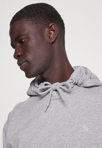 Sweat à capuche gris avec cordon de serrage, texture douce et petit logo blanc sur la poitrine. Il présente une coupe décontractée et des poignets côtelés.
