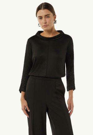 Black long-sleeve top with a cowl neckline, soft texture, and subtle side detailing, paired with black wide-leg pants.
