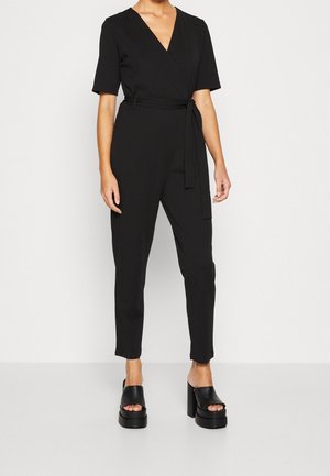 Frau trägt einen schwarzen Kurzarm-Wickel-Jumpsuit mit Taillengürtel und schwarzen Plateau-Sandalen mit offenem Zehenbereich und Absatz, steht vor weißem Hintergrund.