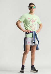 Camiseta atlética verde claro con gráfico rosa, combinada con pantalones cortos grises con acentos rosas. El modelo lleva gafas de sol reflectantes.