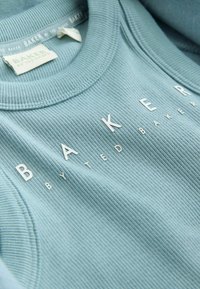 Svetlomodrá rebrovaná tkanina s vystupujúcimi bielymi písmenami tvoriacimi nápis „BAKER BY TED BAKER“ na zloženej mikine s okrúhlym výstrihom.