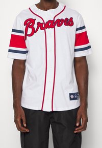 Man draagt een wit Atlanta Braves-shirt met rode en marineblauwe strepen op de mouwen en zwarte korte broek tegen een effen achtergrond.