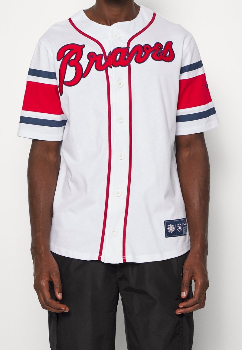 Man draagt een wit Atlanta Braves-shirt met rode en marineblauwe strepen op de mouwen en zwarte korte broek tegen een effen achtergrond.