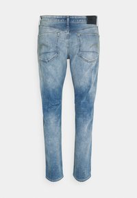 Jean en denim bleu délavé vu de dos avec deux poches et un écusson noir de la marque sur la ceinture.