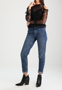 Top in pizzo nero con design off-shoulder e motivi intricati; abbinato a jeans denim blu a vita alta e stivaletti neri con tacco a blocco.