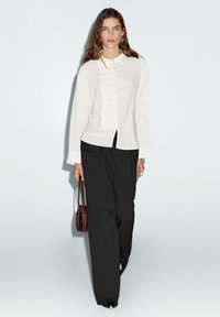 Cremfarbens Button-Up-Hemd mit strukturiertem Vorderteil, kombiniert mit lockeren schwarzen Hosen und einer kleinen braunen Handtasche. Glattes Material, lässige Passform.