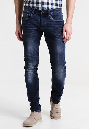 Mies, jolla on tummansiniset slim-fit-farkut, beiget aavikkokengät ja sinivalkoruudullinen paita.