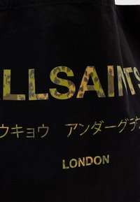 Juodas audeklo laikiklis su didelėmis žaliomis ir gyvūnų rašto užrašais. Dizainas apima 'ALLSAINTS' ir 'LONDON' bei papildomą daugiakalbį tekstą.
