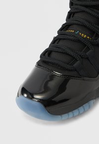 Μαύρο αθλητικό παπούτσι με γυαλιστερό καπάκι στα δάχτυλα, υφασμάτινο πάνω μέρος με υφή και πλεξούδες στα κορδόνια. Διαφανές ελαστικό πέλμα που προσθέτει αντίθεση και ανθεκτικότητα.