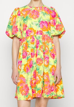 Robe jaune à manches bouffantes ornée de grands motifs floraux roses et orange, portée par une personne debout sur un fond uni.