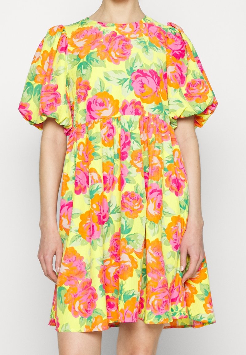 Robe jaune à manches bouffantes ornée de grands motifs floraux roses et orange, portée par une personne debout sur un fond uni.
