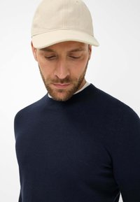 Marineblauer Strickpullover mit rundem Halsausschnitt, kombiniert mit einer hellbeigen Baseballkappe. Die Textur wirkt glatt und hat eine taillierte Passform.