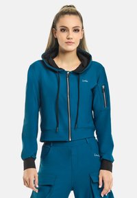 Korte teal hoodie met zwarte voering, voorrits, verstelbare koordjes en een ritszak op de linkermouw. Glad textuur van de stof.