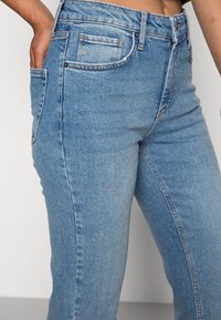 Närbild av en person som har ljusblå denimjeans och en hand i bakfickan framför en enfärgad bakgrund.