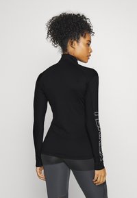 Schwarzes Langarmoberteil mit hohem Kragen, gerippter Textur und einem Logo, das auf dem linken Ärmel aufgedruckt ist. Das Material scheint flexibel und enganliegend zu sein.