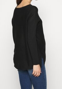 Svart stickad tröja med långa ärmar, med ribbad textur och sidoklipp, tillsammans med blå jeans.