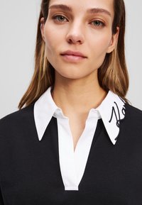 Svart polo skjorte med hvit krage, med broderte tekstdetaljer på kragen, som fremhever et strukturert design og teksturert stoff.
