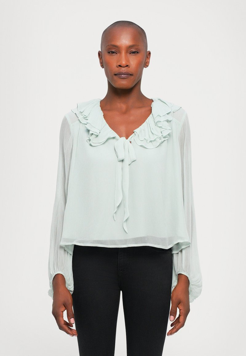 Light mint green blouse with ruffled neckline and tie detail. Long, gathered sleeves and a loose fit, made from a lightweight, textured fabric.