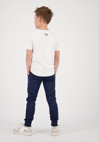 T-shirt blanc à manches courtes avec un logo noir dans le dos, associé à un pantalon de jogging marine et des baskets blanches. Texture lisse, design décontracté.