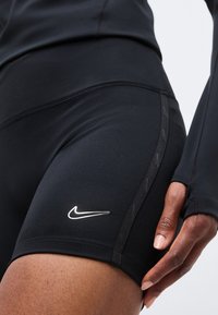 Κοντινό πλάνο ατόμου που φοράει μαύρα αθλητικά σορτς Nike και μαύρο μακρυμάνικο τοπ, το χέρι ξεκουράζεται κοντά στον μηρό, εμφανίζοντας το λογότυπο Nike στα σορτς.