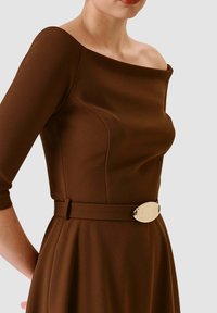 Vestito marrone con scollo a barca, corpetto aderente e maniche a tre quarti. Presenta un dettaglio decorativo con fibbia argentata sulla cintura.