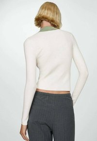 Pull court à manches longues blanc avec un col vert, présentant une texture lisse et une silhouette ajustée, associé à un pantalon gris à fines rayures.