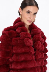 Palton roșu din blană artificială, cu segmente pufoase și suprapuse. Textură moale, guler larg și un aspect luxos. Are un design proeminent, rotunjit.