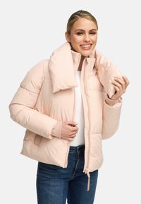 Lächelnde Frau, die eine hellrosa Daunenjacke mit großem Kragen, ein weißes Hemd und blaue Jeans trägt und mit der rechten Hand den Kragen anpasst.