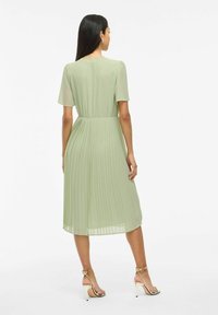 Femme portant une robe midi plissée vert clair avec des manches courtes et des sandales à talons beiges, se tenant de dos par rapport à l'objectif.