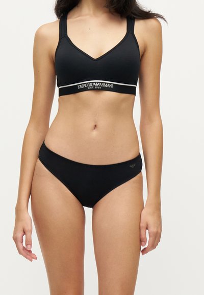 Femme portant un soutien-gorge noir Emporio Armani et une culotte bikini noire assortie sur fond blanc uni.