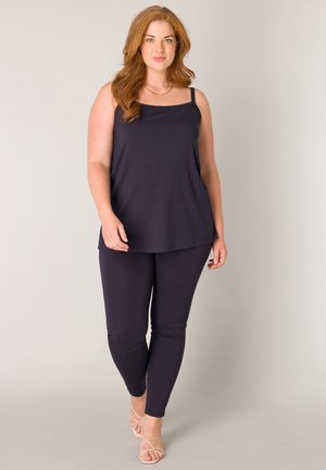 Vrouw met rood haar, gekleed in een zwart mouwloos topje, zwarte legging en beige sandalen met bandjes, staand tegen een effen achtergrond.