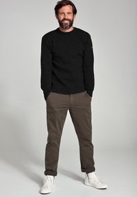 Un pull noir à col rond, un pantalon tapered vert olive et des baskets blanches. La tenue est décontractée et présente une coupe ample.