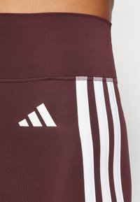 Burgunderry idrettsleggings med en glatt tekstur, med tre hvite striper langs siden og en hvit logodetalj.