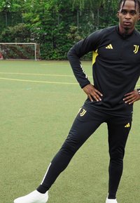 Schwarzer, sportlicher Trainingsanzug mit gelben Akzenten, bestehend aus einer zip-up Jacke und eng anliegenden Hosen. Trägt weiße Schuhe und steht auf einem grünen Feld.