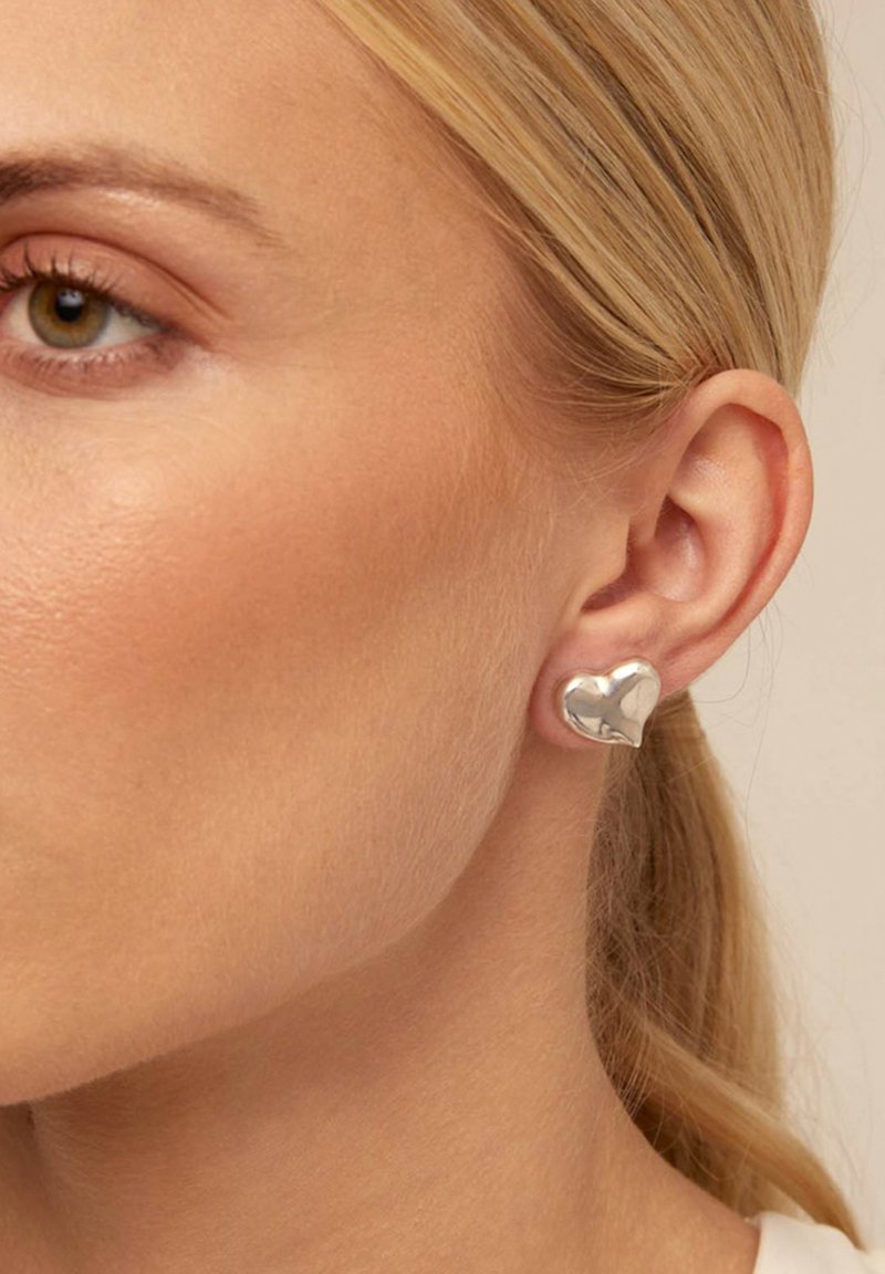 Pendiente plateado en forma de corazón con una superficie lisa y reflectante. El diseño es minimalista y contemporáneo, ajustándose cómodamente contra la oreja.
