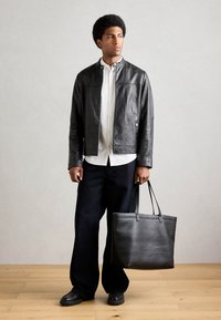 Schwarze Lederjacke mit Druckknopfkragen, getragen über einem weißen Hemd und dunklen, weiten Jeans. Hält eine große schwarze Lederhandtasche.