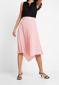 Vaaleanpunainen pleatsihame, jossa on korkea-matala helma ja tiukka vyötärö. Yhdistetty mustan ilmanhihaisen paidan ja mustien hihnalokareiden kanssa.