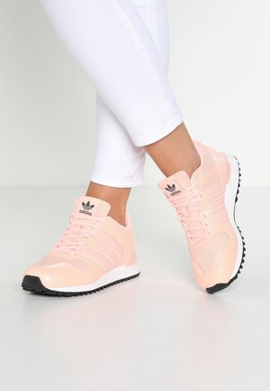 Pieds chaussés de baskets Adidas rose pâle avec semelles blanches et crampons noirs, assorties à un pantalon blanc ajusté sur un fond clair uni.