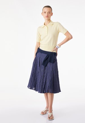 Nainen seisoo käsi lanteilla, pukeutuneena vaaleankeltaiseen lyhythihaiseen poolopaitaan, tummansiniseen ruudulliseen midi-hameeseen, valkoisiin sandaaleihin ja vyötärölle solmittuun tummansiniseen neuleeseen.