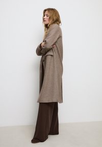 Cappotto a scacchi marrone con vestibilità rilassata, maniche lunghe e chiusura doppiopetto, abbinato a pantaloni a gamba larga marrone scuro.