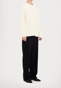 Pull en maille côtelée crème à col rond, assorti d'un pantalon noir ample et de chaussures noires pointues, présenté sur un fond neutre.