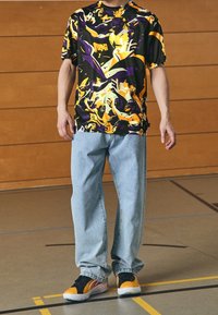 Svart, lila och gul abstrakt mönstrad t-shirt; ljusblå denimjeans; svarta och gula sneakers med vita sulor.