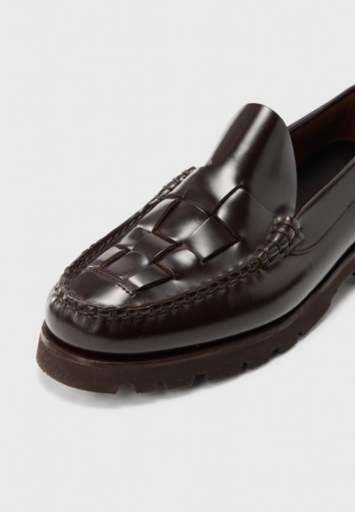 Pantof din piele maro închis, cu detaliu împletit pe curea și talpă groasă de cauciuc, fotografiat pe un fundal simplu, deschis la culoare.