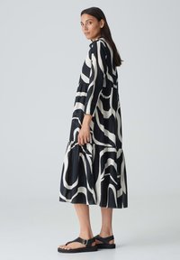 Schwarz-weißes, gestuftes Kleid mit Wellenmuster, langen Ärmeln und einem lässigen Schnitt. Kombiniert mit flachen schwarzen Sandalen.