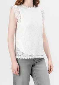 Vit spetsblus med scallopkanter och korta ärmar, prydd med blommönster och semi-transparent struktur. Tillsammans med grå jeans.