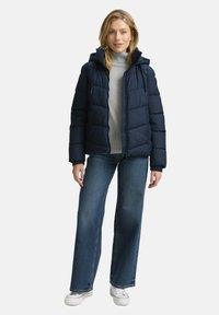 Dunkelblaue Steppjacke mit Kapuze, die gesteppten Abschnitte aufweist. Getragen über einem grauen Pullover und kombiniert mit weit geschnittenen Jeans und weißen Sneakers.